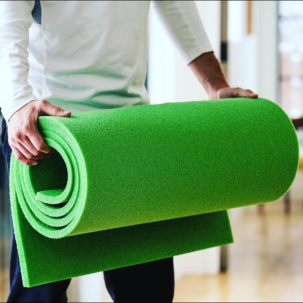 A person in a white long-sleeve shirt holds a large, rolled-up BirdieBall Indoor Putting Green - Classic Depth Putting Mat indoors.