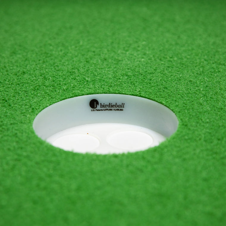 A close-up of the BirdieBall Double Depth Putting Green Cup (1" Deep), featuring the logo, set in lush green artificial turf.