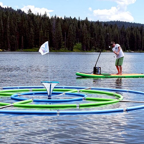 In the BirdieBall Target Golf Game, a man on a paddleboard tries to land shots on a floating target with rings and a flag, set on a scenic lake surrounded by trees and hills.